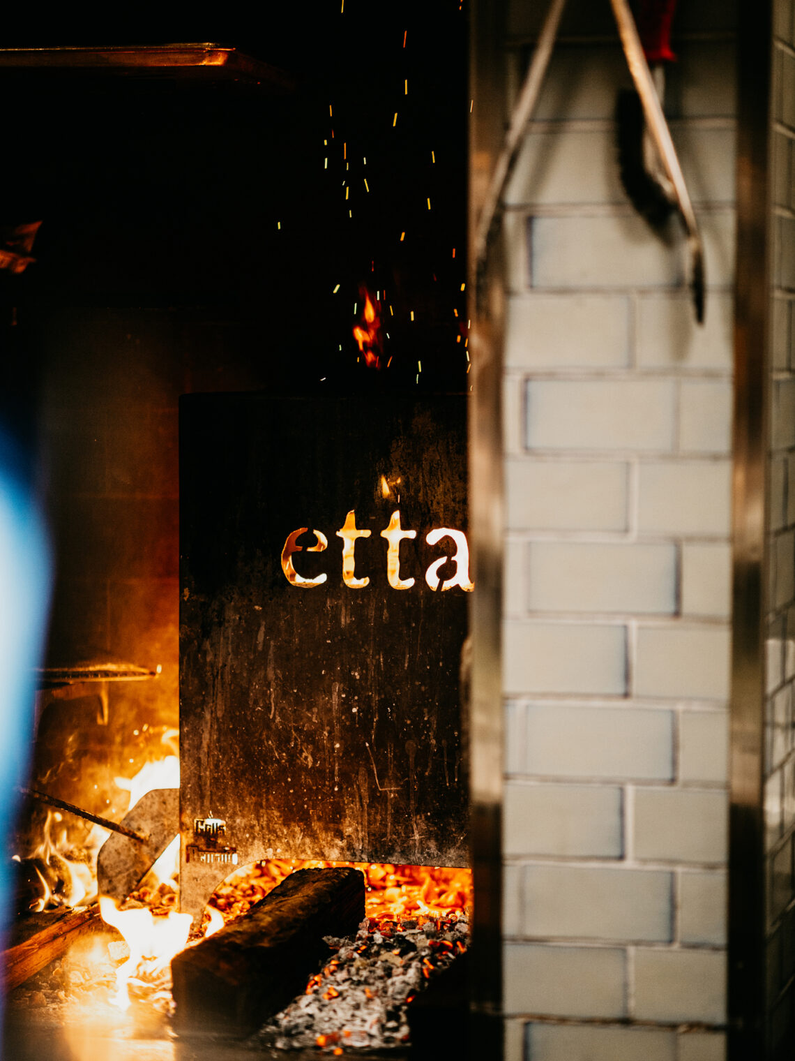 Wood fire burning in a restaurant hearth featuring a metal plate cutout spelling "etta".