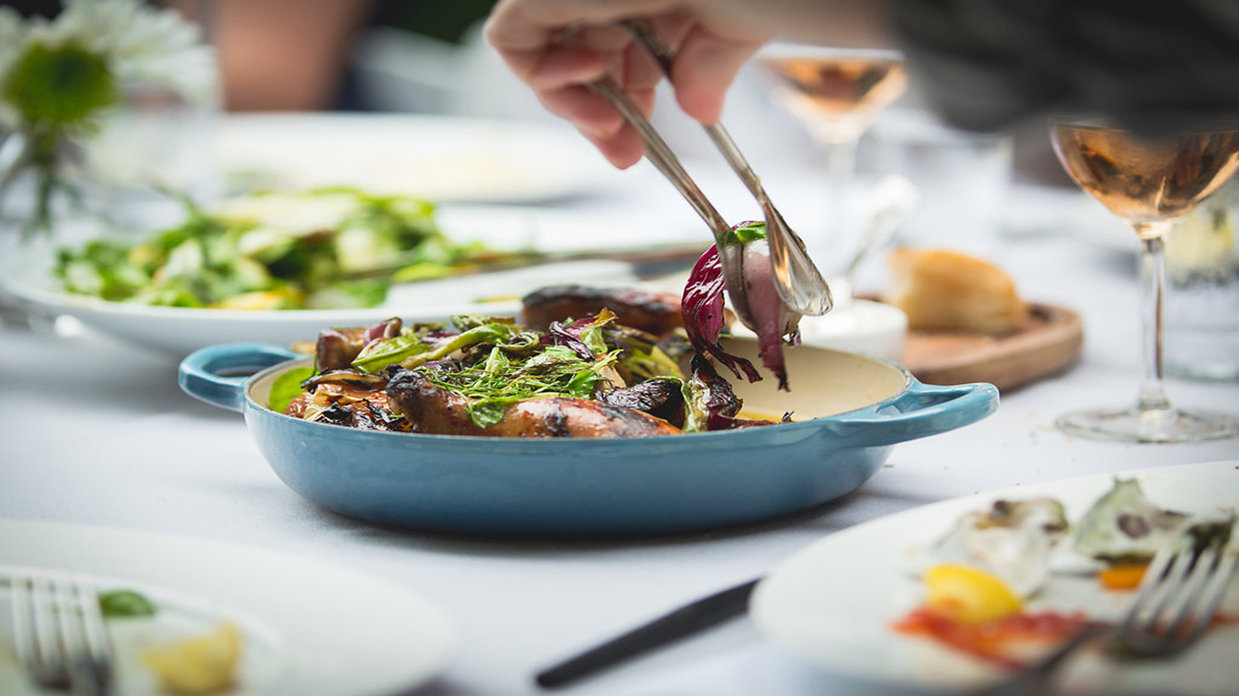 Tongs serving a roasted vegetable dish from a blue ceramic pan on a restaurant table.