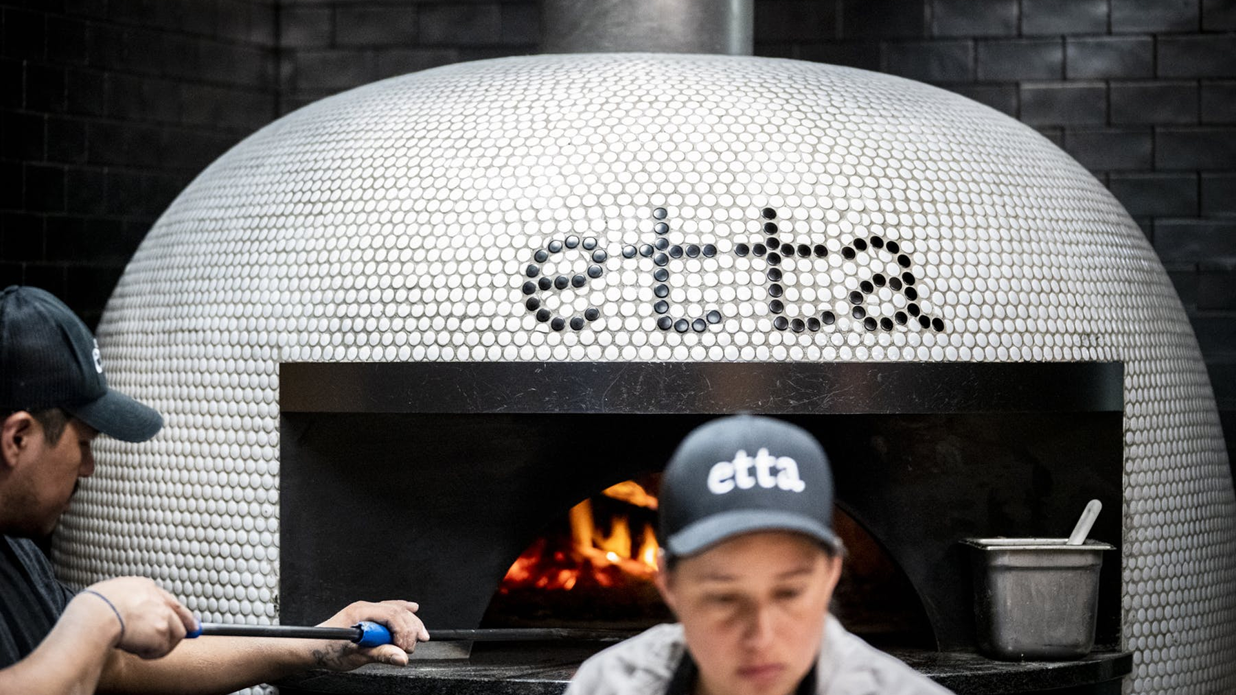 Chefs working in front of a white tiled wood-fired pizza oven with "etta" logo.