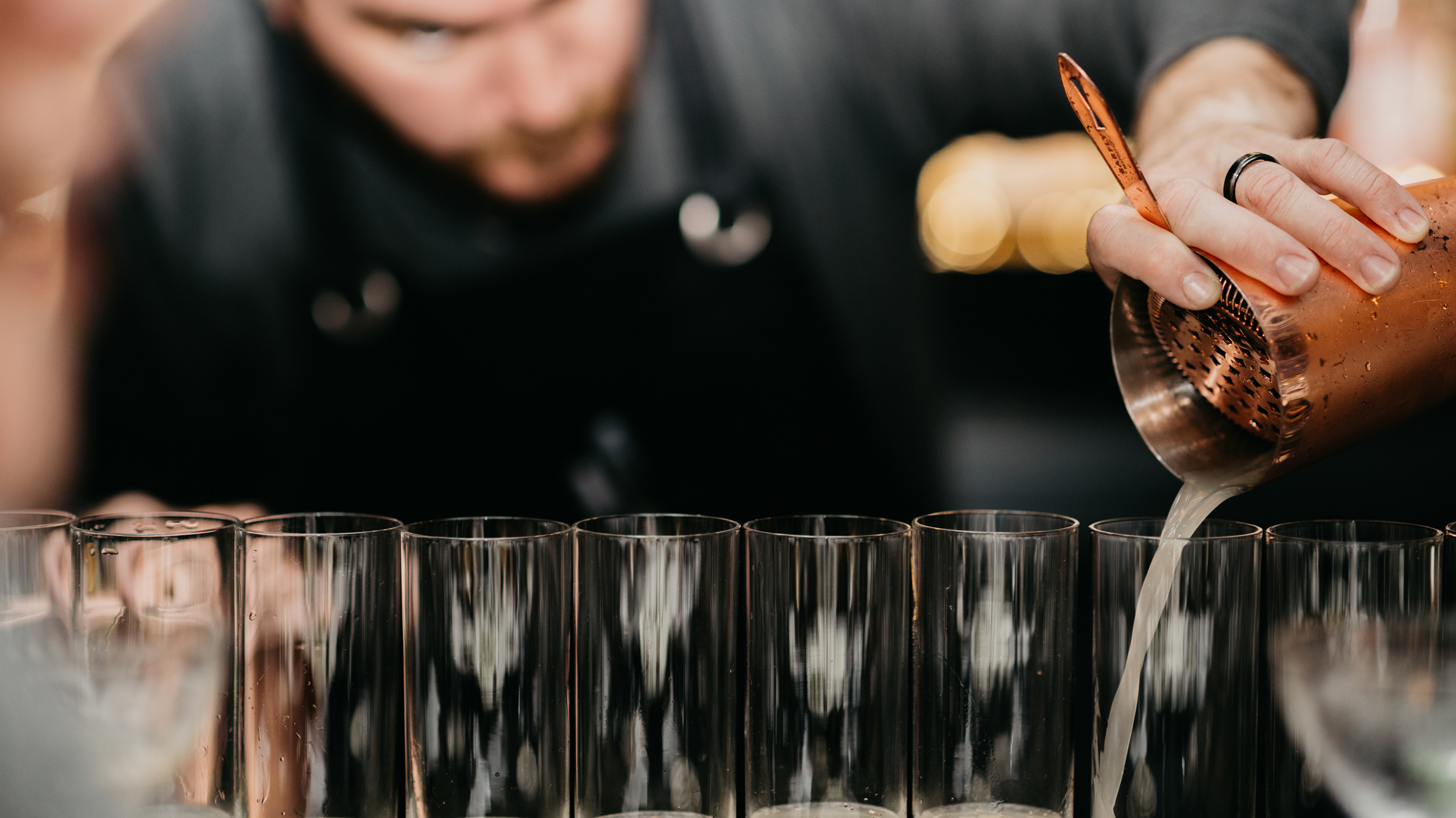 Close-up of a bartender pouring a cocktail from a copper shaker into tall glasses.