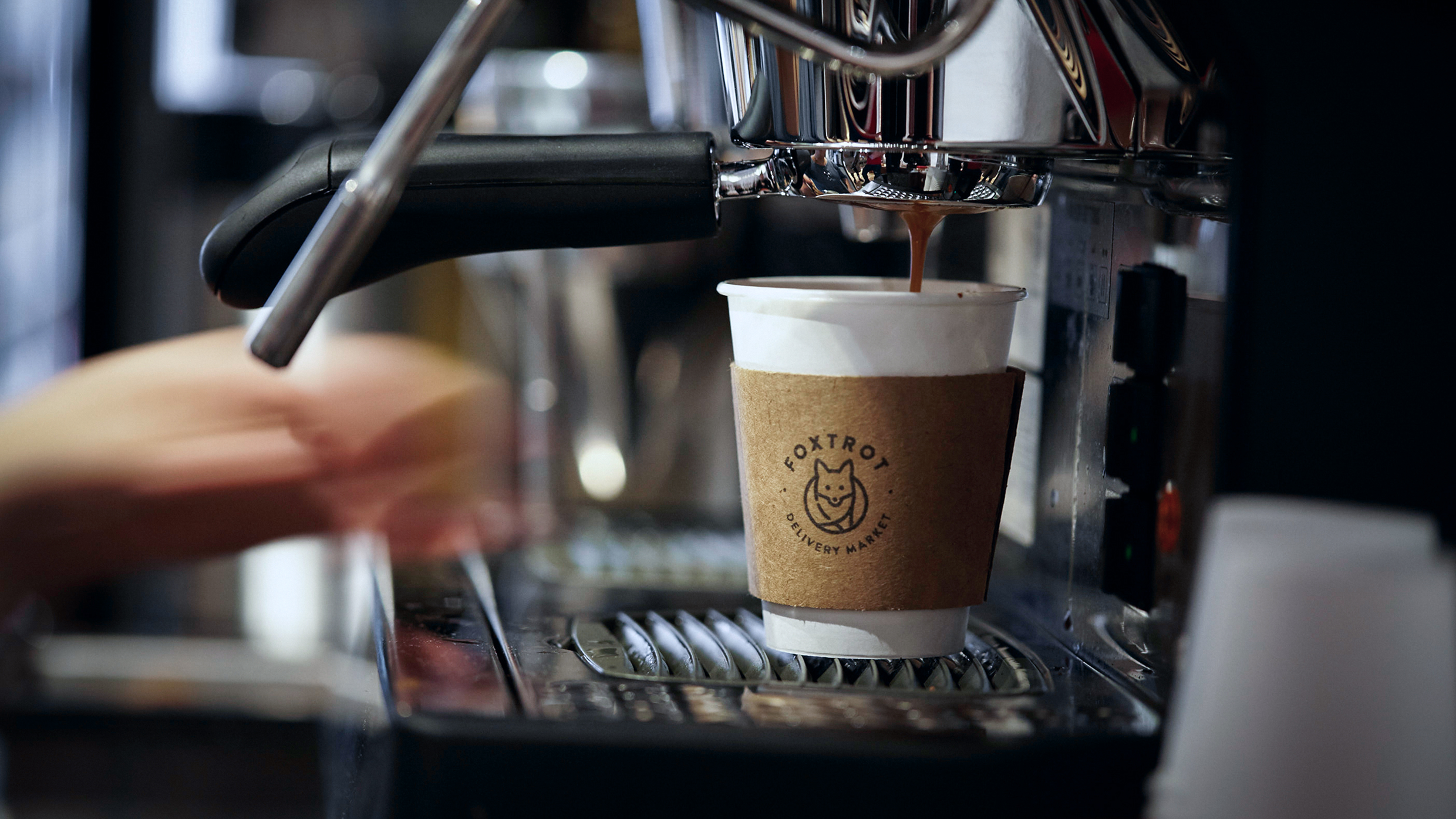 A white paper coffee cup with a Foxtrot branded sleeve under an espresso machine.