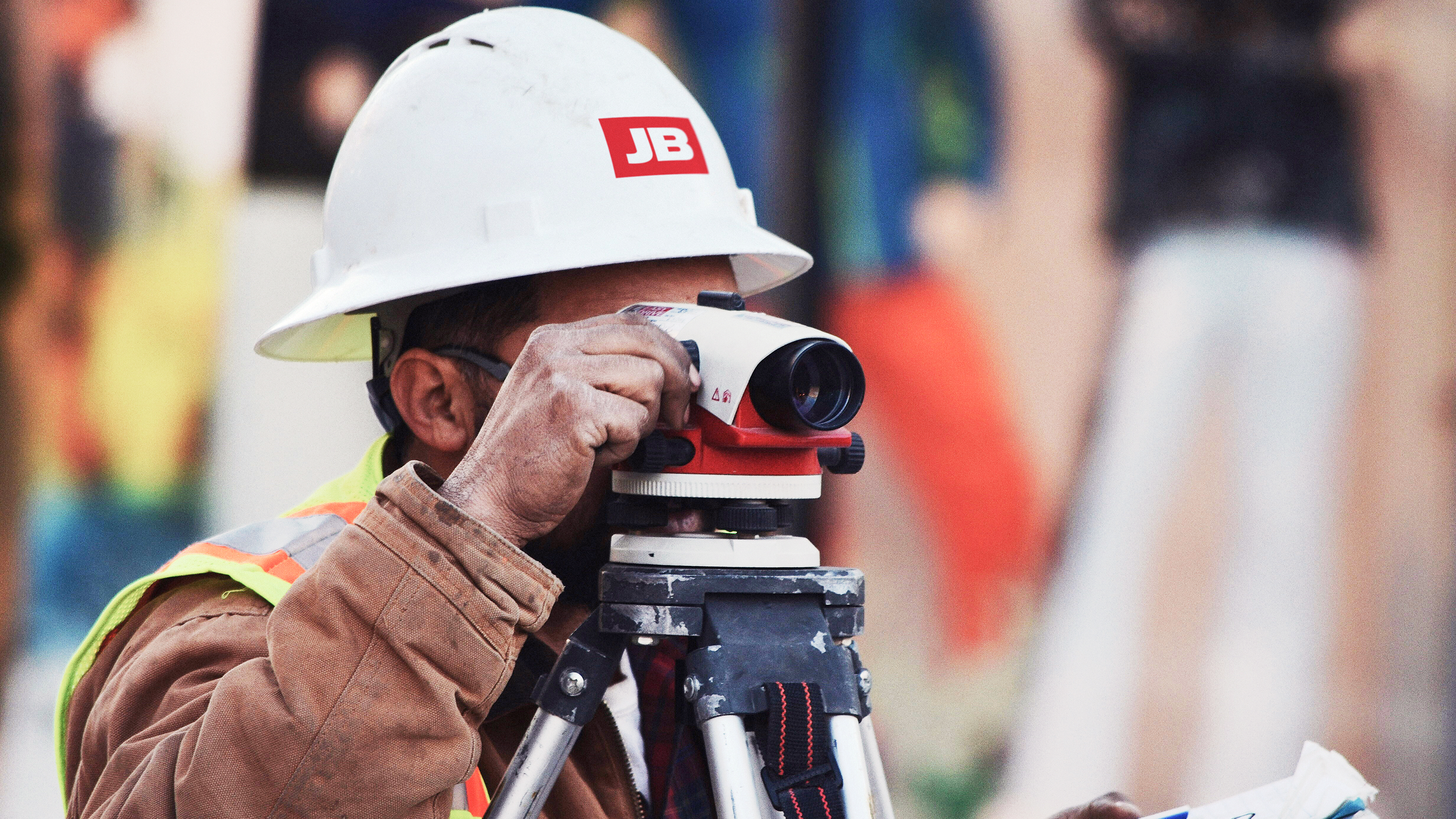 Construction worker wearing a JB hardhat looking through a land surveying transit level.