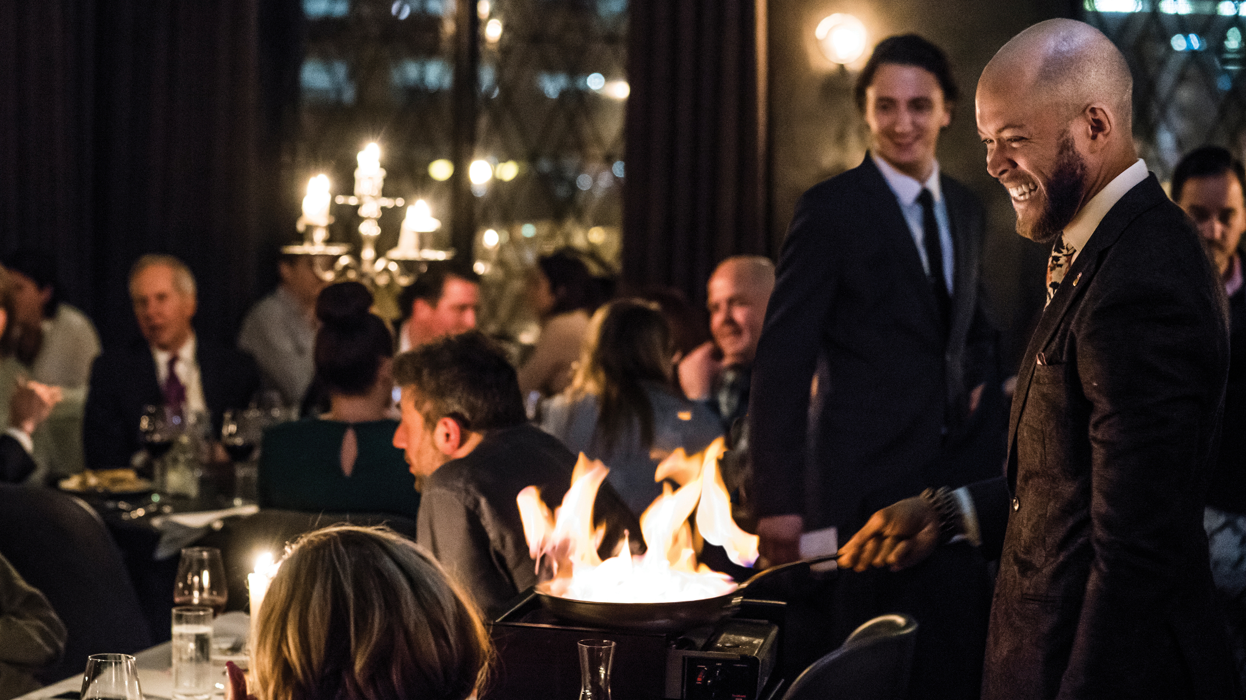 A smiling chef flambés a dish tableside in a dimly lit, elegant restaurant filled with diners. The flames from the pan illuminate the scene, creating a lively and sophisticated atmosphere.