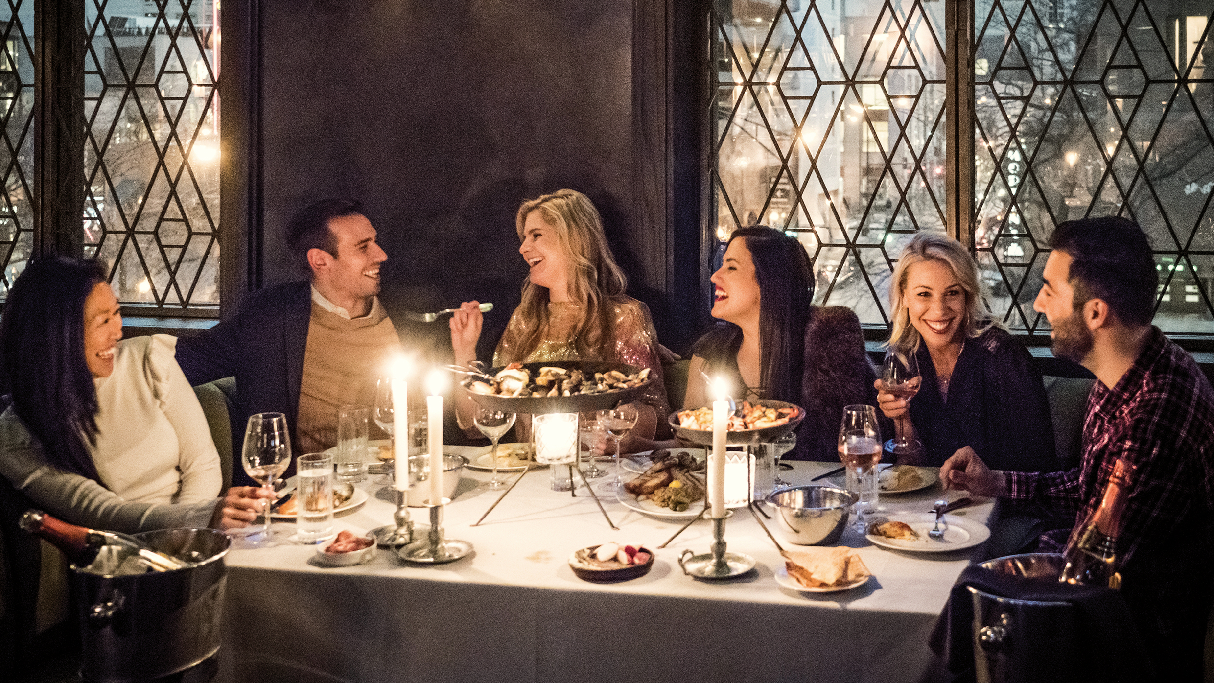 A group of six people laugh and enjoy dinner together at a candlelit restaurant table, with large windows and platters of food and drinks in front of them.