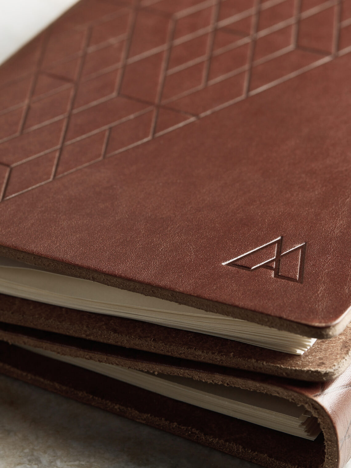 Close-up of two brown leather-bound notebooks stacked, featuring geometric patterns and an embossed logo on the cover, with slightly textured paper edges visible.