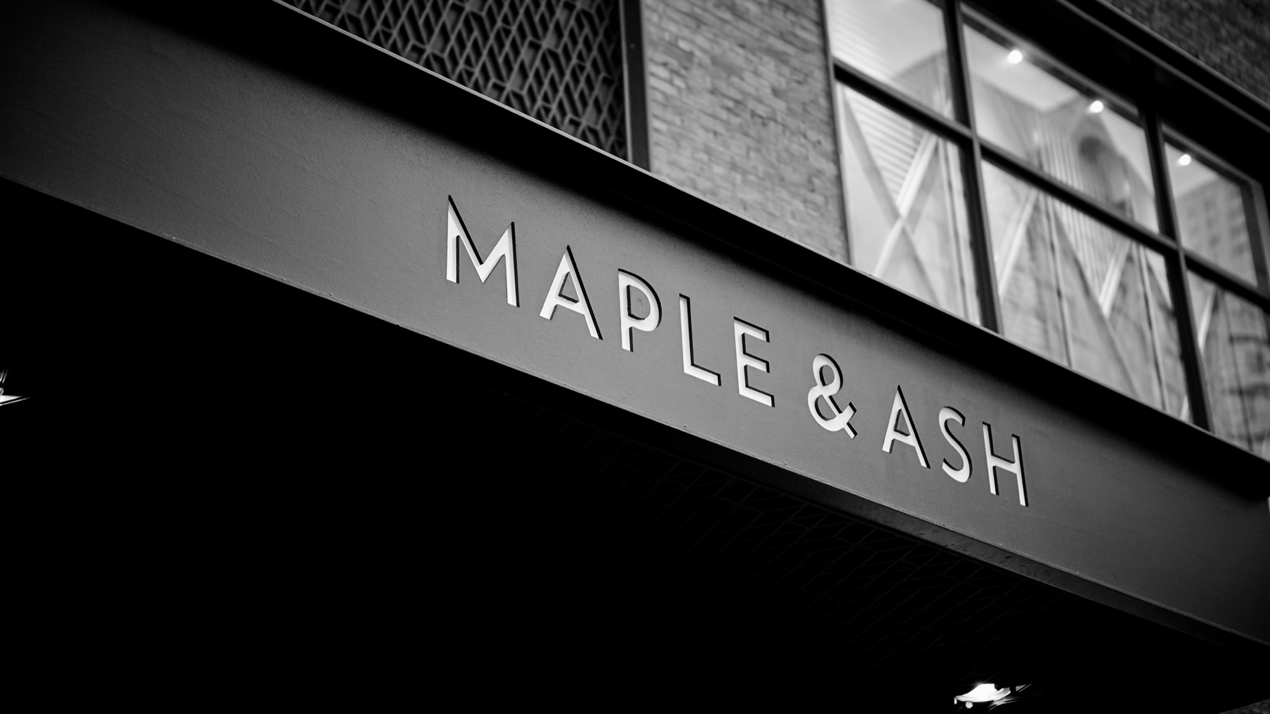 Black and white photo of a building awning displaying the restaurant name MAPLE & ASH in large, bold letters, with windows and brickwork visible above.
