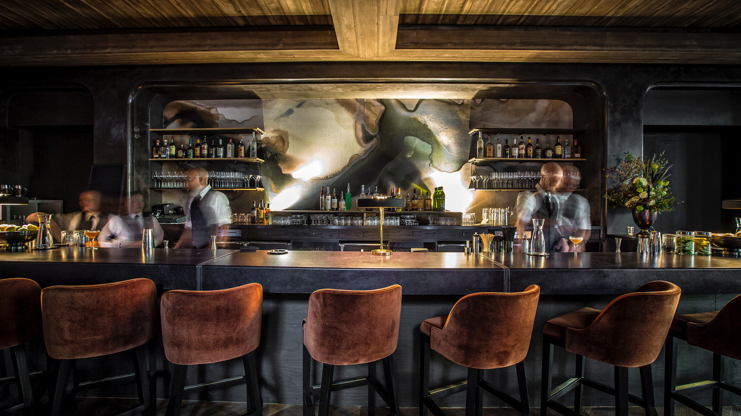 A modern bar with five velvet barstools, a dark counter, and shelves of bottles behind. Four blurred bartenders work behind the bar, with warm lighting and abstract wall art creating a stylish, inviting atmosphere.
