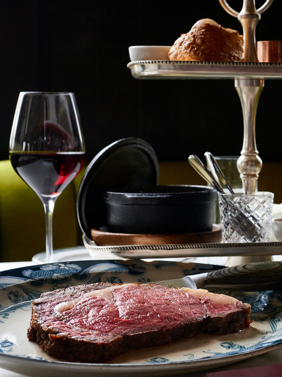 A thick slice of rare roast beef on a patterned plate, next to a glass of red wine, with a bread roll and condiments on a tiered stand in the background.