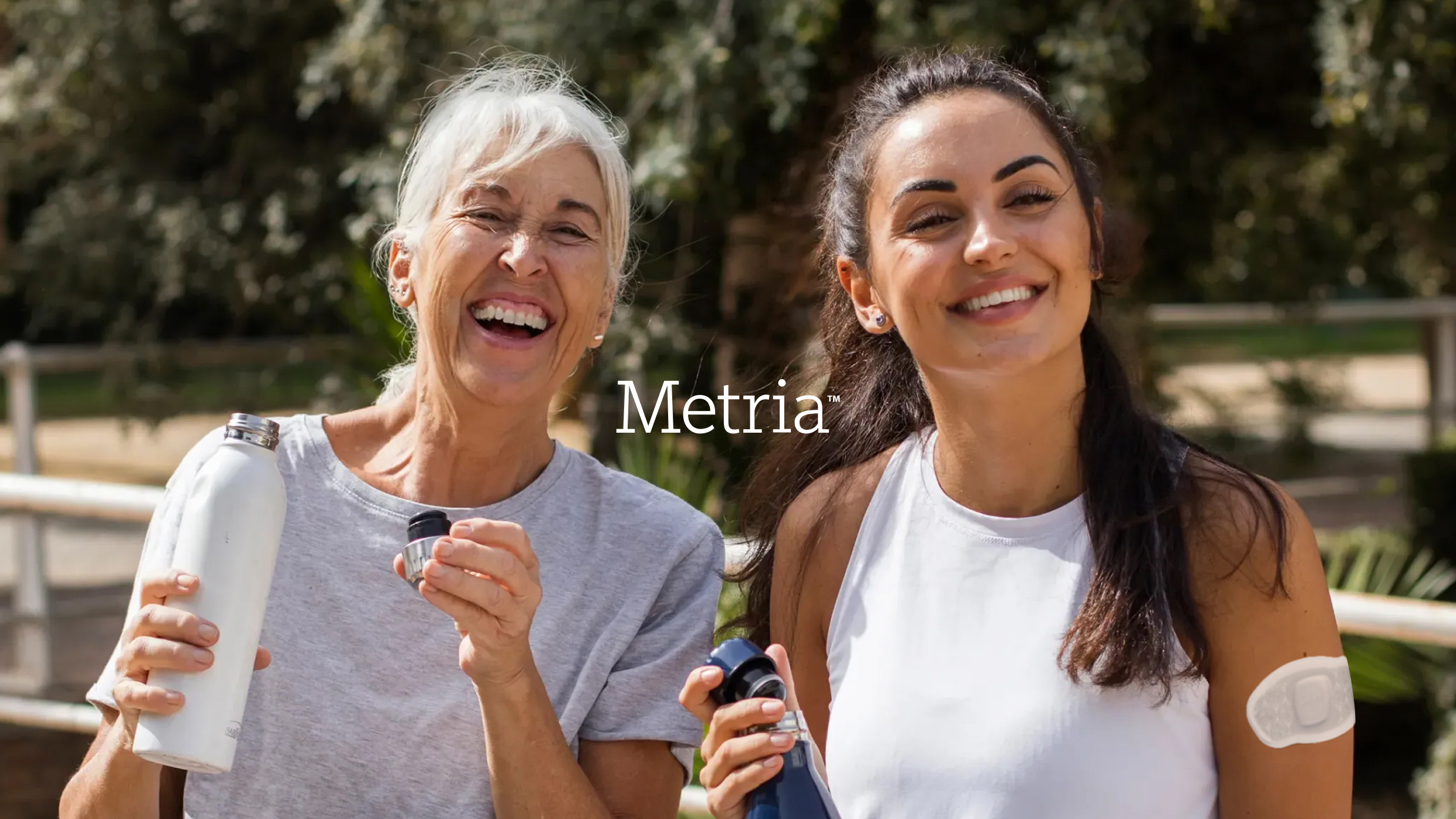 Two smiling women holding water bottles, with one wearing a Metria health patch.