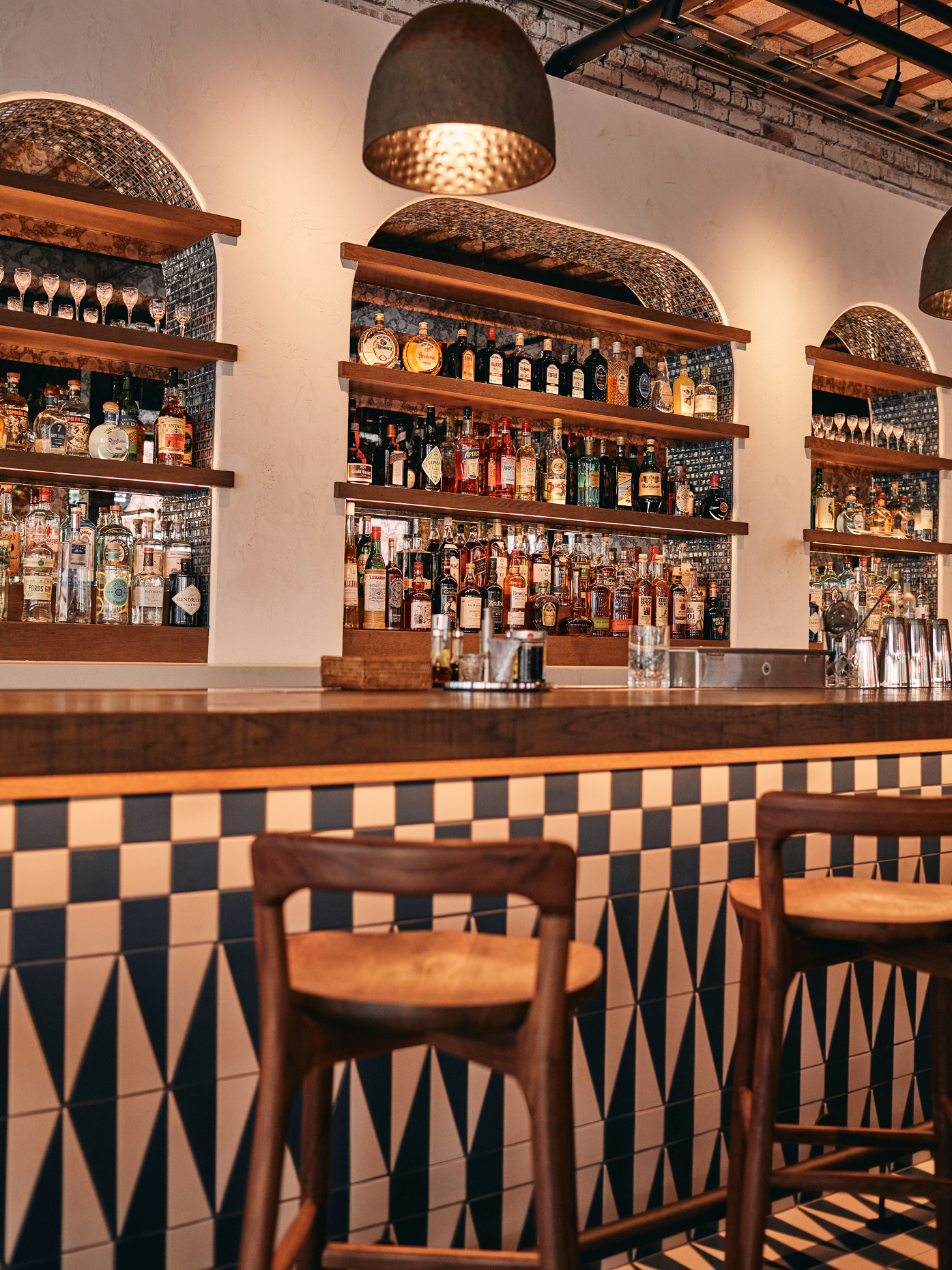 A stylish bar with a patterned tile counter, wooden chairs, and shelves stocked with various bottles of liquor and glassware under arched alcoves, illuminated by warm pendant lights.
