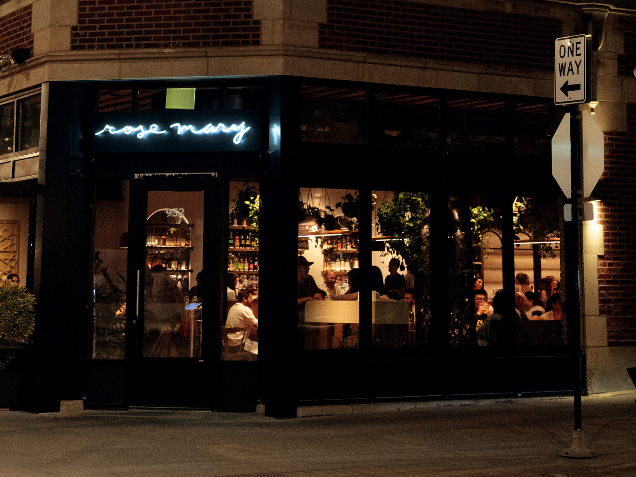 A warmly lit restaurant called Rose Mary with large front windows showing people dining inside at night; a neon sign is above the entrance and a one way street sign is visible on the corner.