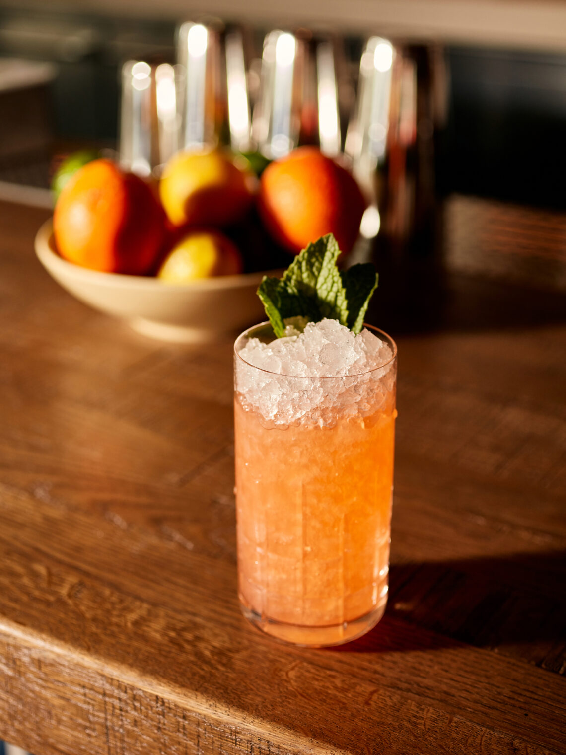A tall glass of orange-pink cocktail topped with crushed ice and mint leaves sits on a wooden bar. In the background, a bowl of citrus fruits and metal shakers are visible.