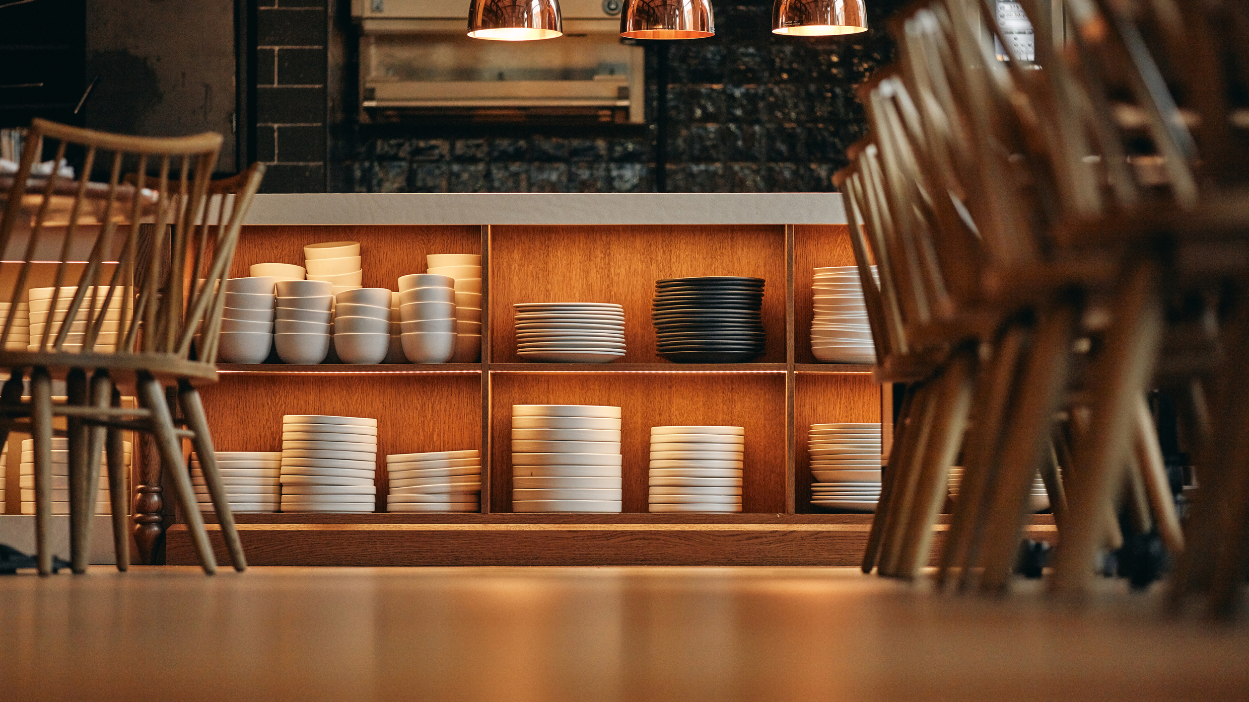 Wooden shelves filled with neatly stacked white and black plates and bowls are shown in a warmly lit restaurant interior, with wooden chairs placed around tables in the foreground.
