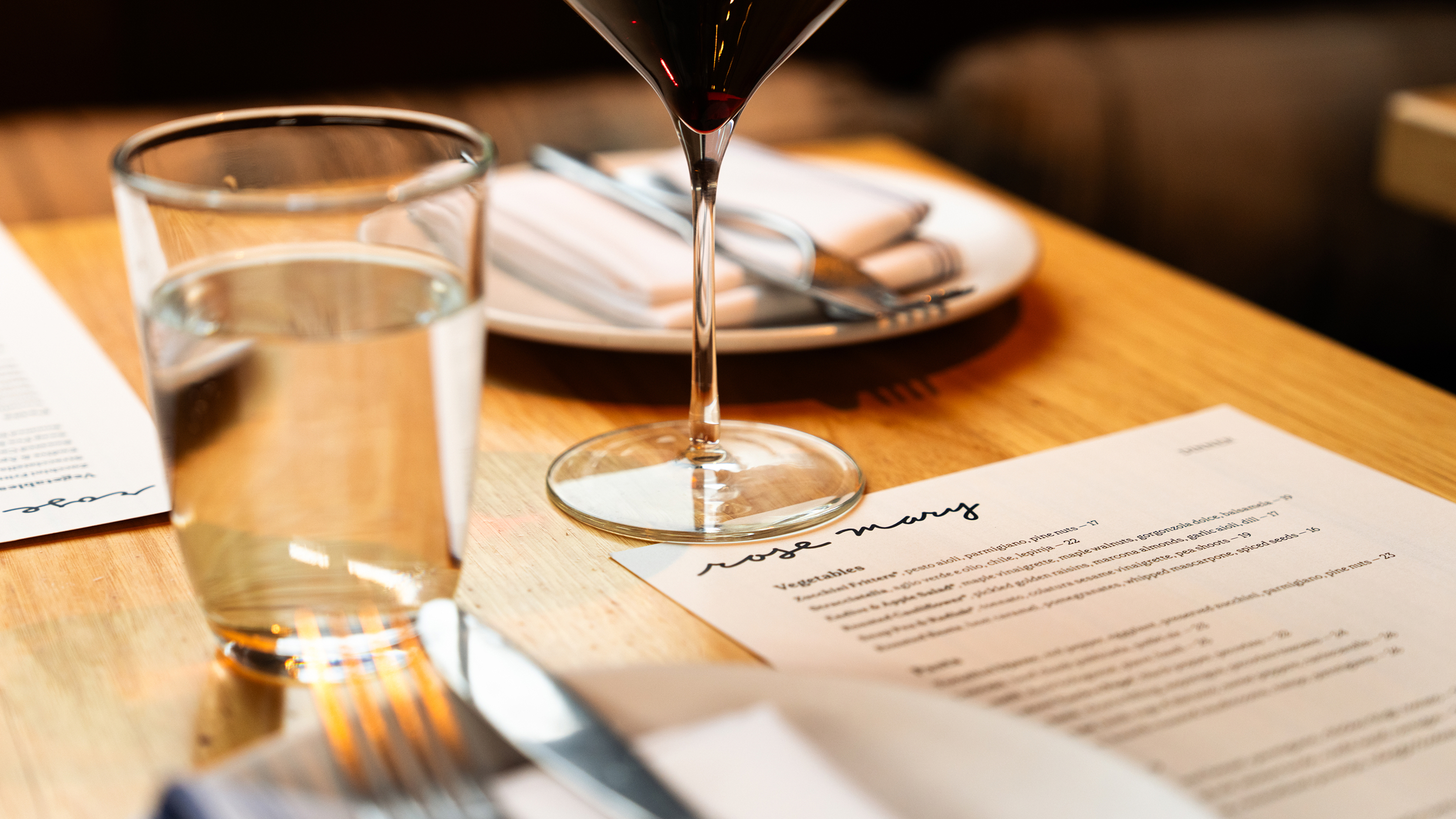 A close-up of a restaurant table set with a glass of water, a glass of red wine, a plate with utensils, and a printed menu on a wooden surface. Soft lighting creates a warm, inviting atmosphere.