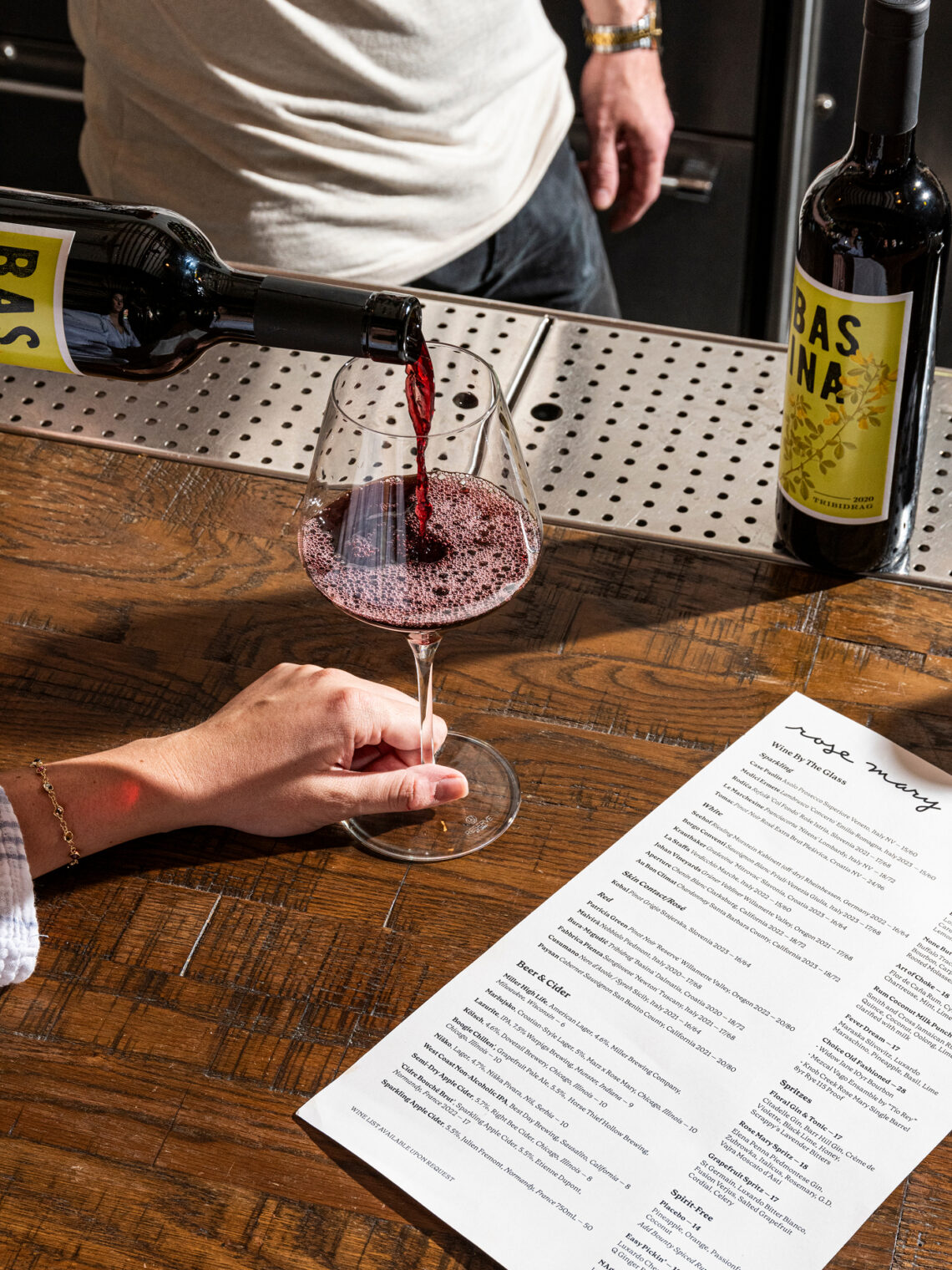 Pouring red wine into a glass next to a Rose Mary drink menu.