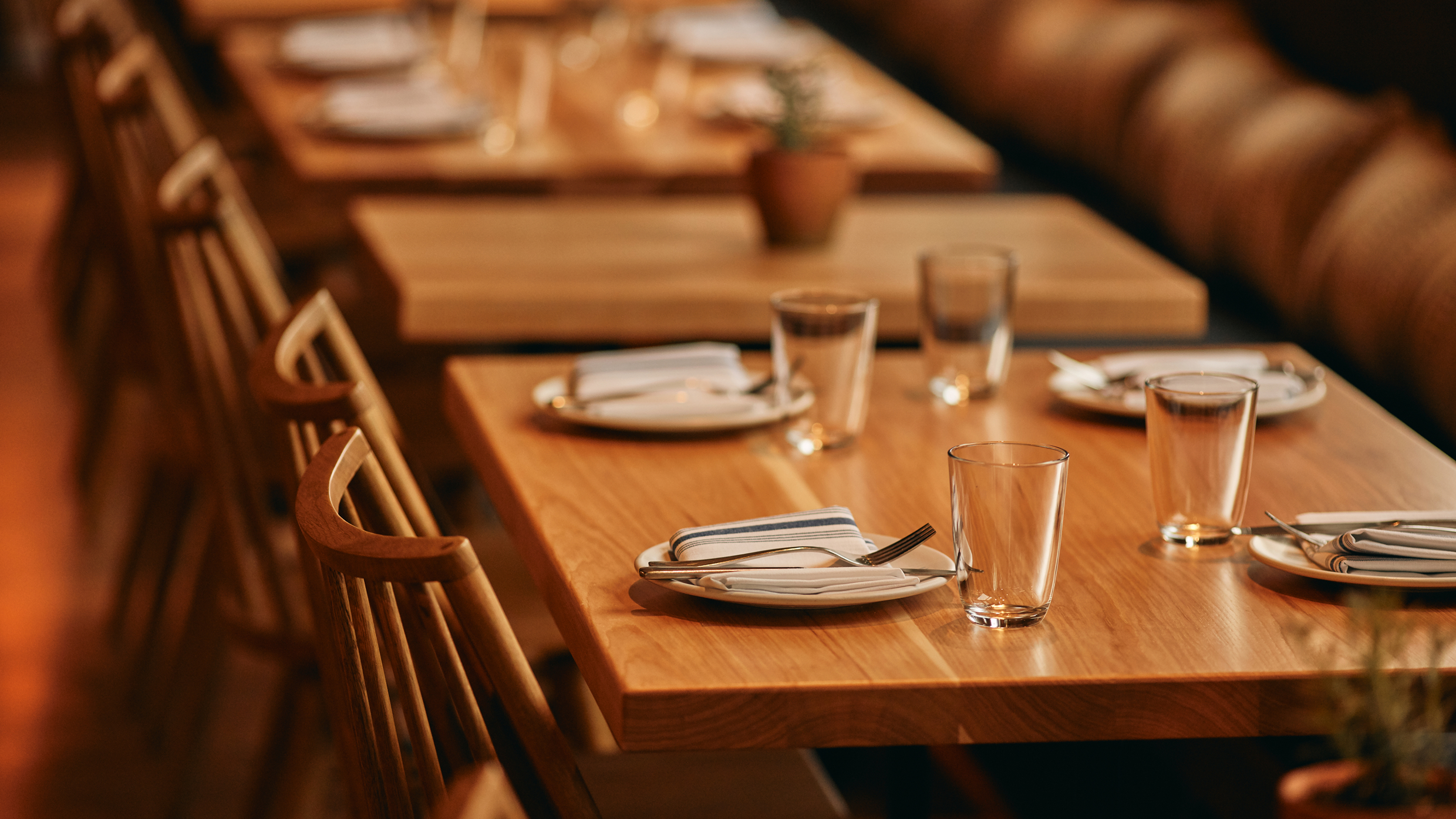 Wooden tables set with plates, napkins, cutlery, and glasses are arranged in a cozy restaurant. The focus is on the neatly set tables, with soft, warm lighting and blurred background seating.