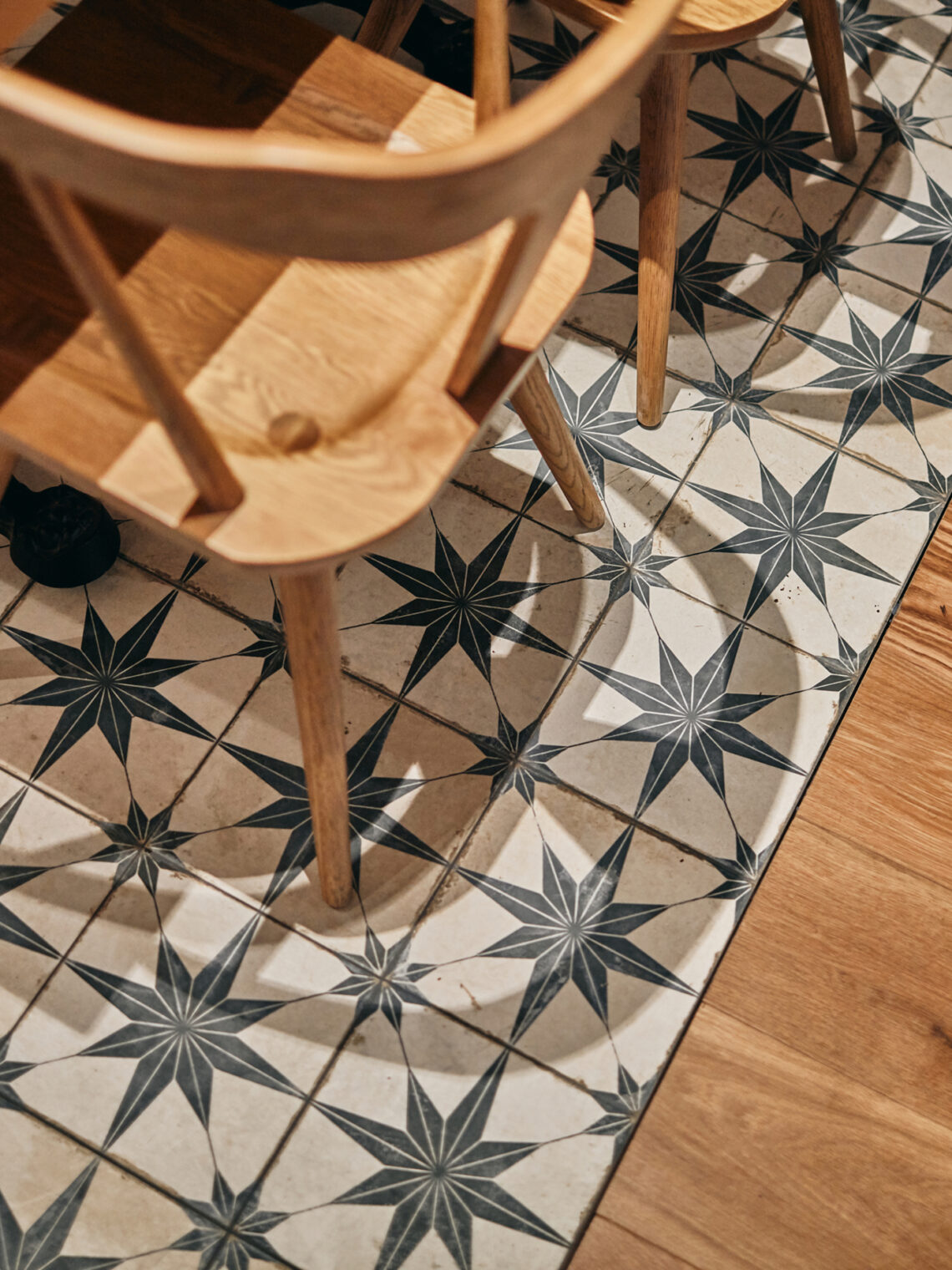 A wooden chair sits on a patterned tile floor with black star designs, adjacent to a wooden floor. The image captures part of the chair and the geometric contrast between the two flooring types.