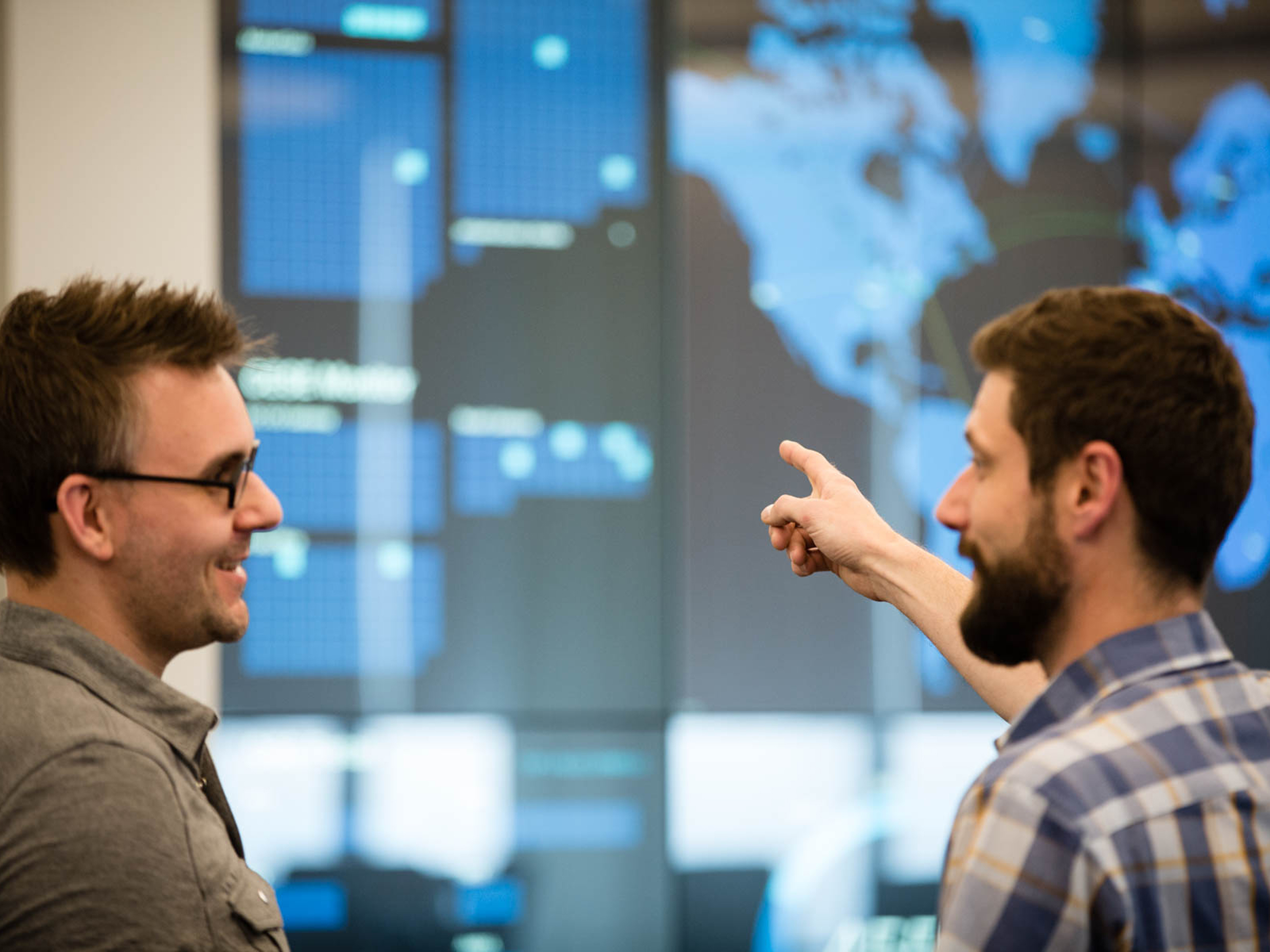 Two men in casual office attire discuss data while standing in front of a large digital display showing global maps and analytics screens.